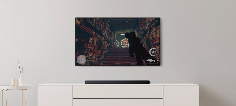 In einem Wohnzimmer befinden sich ein TV und eine Soundbar. Auf dem TV-Bildschirm läuft ein FPS-Spiel und der TV-Kanal ist auf Fußballspiel eingestellt (Video abspielen).