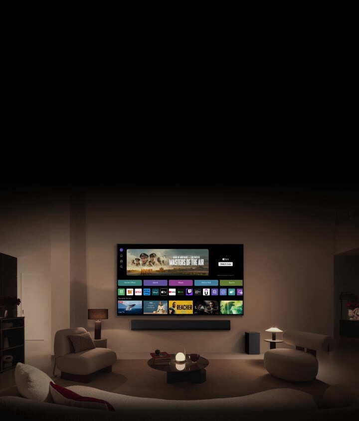 LG OLED TV showing the buttons Home Office, Game, and Music over a banner for Masters of the Air zooms out to show the TV mounted on a wall in a living room. The following logos are displayed on the TV screen in the image: LG Channels, Netflix, Prime Video, Disney TV, Apple TV, YouTube, Spotify, Twitch, GeForce Now and Udemy.