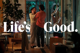 Ein lächelndes Paar steht in einem warm beleuchteten, modernen Wohnzimmer mit bodentiefen Fenstern und Zimmerpflanzen. Großformatiger weißer Text 'Life’s Good.' ist eingeblendet.