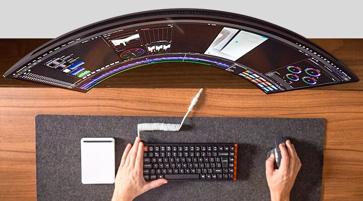 Person arbeitet am Schreibtisch, Hände bedienen Maus und Tastatur vor einem breiten Monitor.