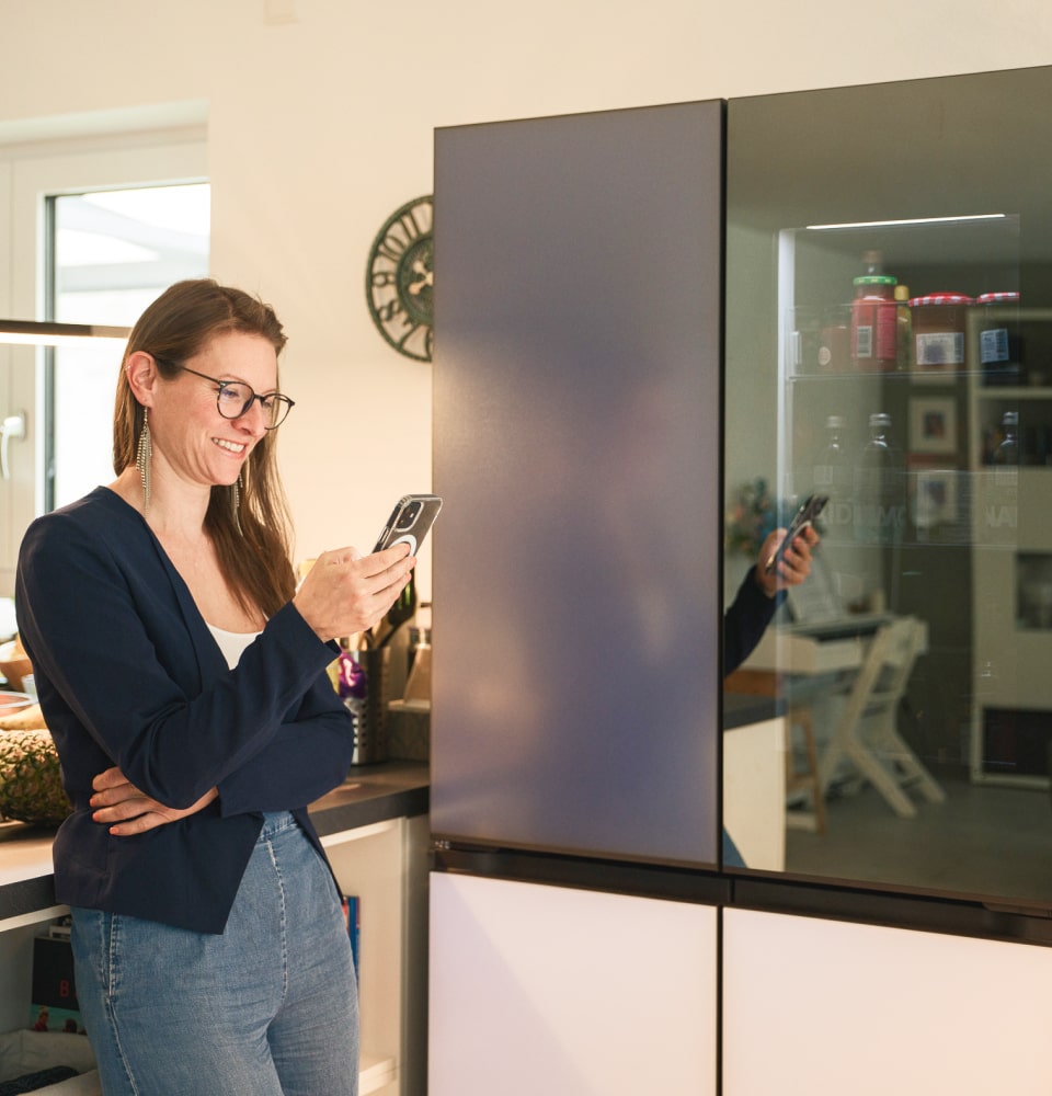 Eine Frau benutzt die ThinQ-App auf ihrem Telefon, um Musik über Bluetooth vor dem LG MoodUP Refrigerator abzuspielen.