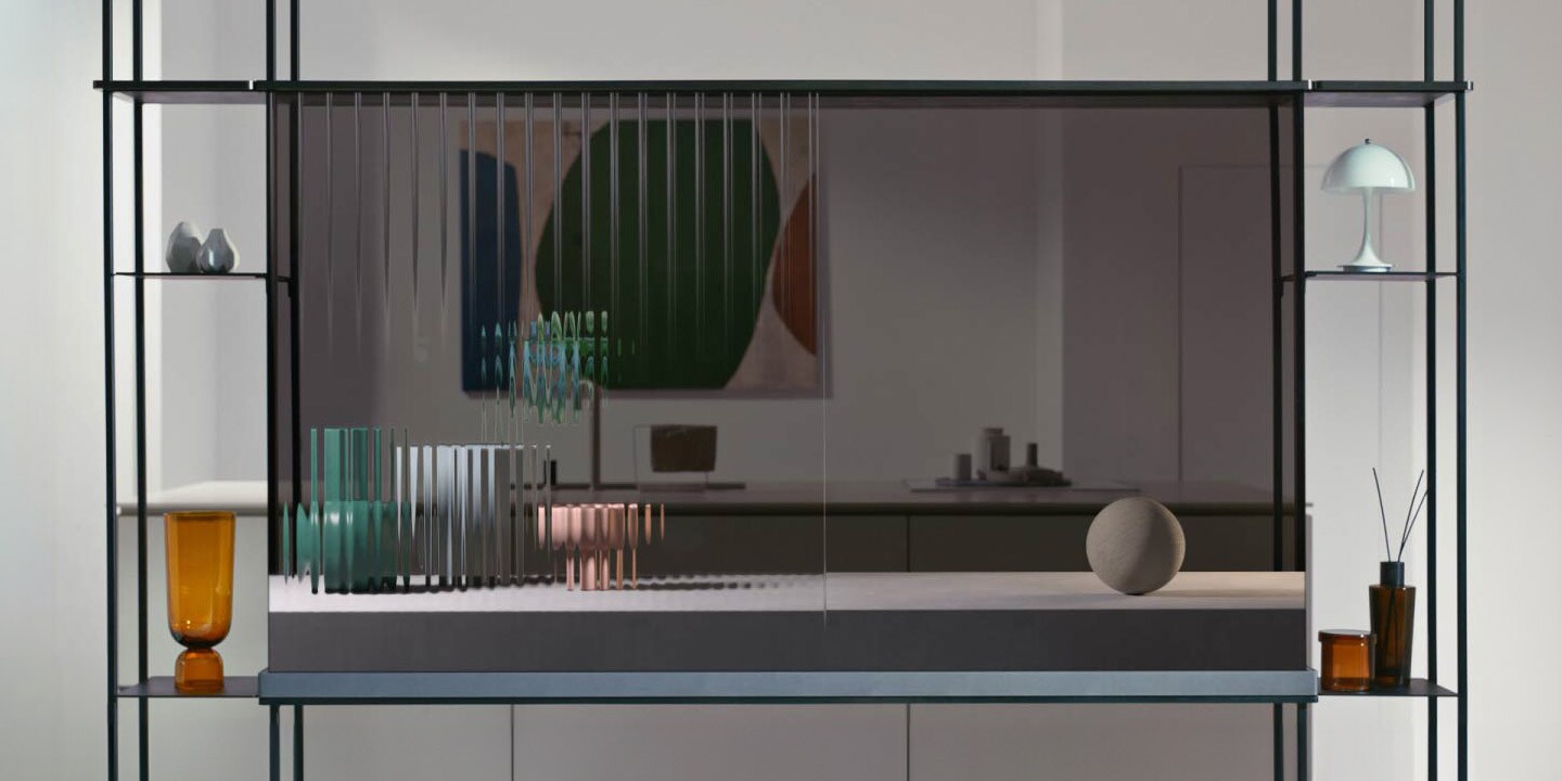 Der LG SIGNATURE OLED T zeigt eine Katze, die über den transparenten Bildschirm springt, während er gleichzeitig den dahinter liegenden Innenraum zeigt. Rechts und links des modularen Regals sind Gegenstände angebracht. Ein weiterer transparenter Fernseher steht vor dem Fenster, das die Winterlandschaft außerhalb des Hauses einrahmt. Auf dem Bildschirm sind fallende Blütenblätter zu sehen. Der LG SIGNATURE OLED T aus der ersten Szene zeigt nun drei Töpferobjekte, die sich von unscharfen Linien zu einem verfestigten Objekt entwickeln.