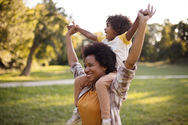 Afroamerikanische Mutter und Tochter stehen in einem Park. Kleines Mädchen auf den Schultern der Mutter