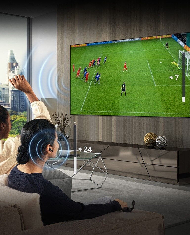 A group of people are sitting on a sofa watching a soccer game on TV. The woman on the far right is wearing earbuds and using them with a different volume than the TV, indicating that she is using both at the same time.