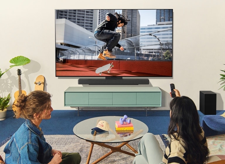 سيدتان تجلسان على أريكة أمام LG TV مثبت على الحائط في غرفة معيشة ملونة، وإحداهما تشير إلى جهاز التحكم عن بعد إلى شاشة التلفزيون. 