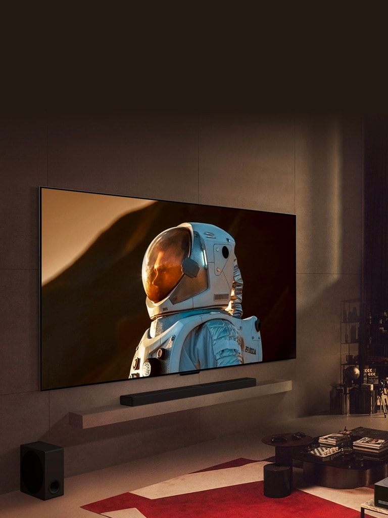 LG TV كبير جدًا مثبت على الحائط في غرفة معيشة مريحة ويعرض لقطة قريبة لرائد فضاء، ويشغل معظم مساحة الجدار. رجل يجلس على أريكة ويشاهد التلفاز أمامه مع كلب، مع عرض منظر المدينة ليلاً خلف النافذة.
