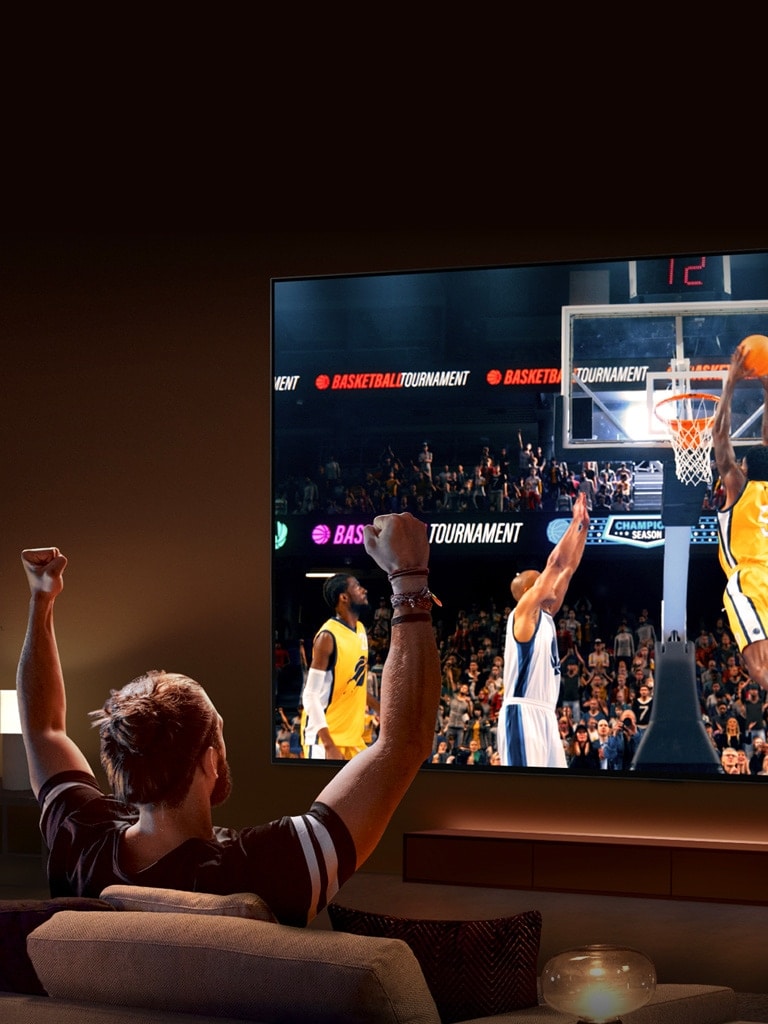 رجل يجلس على أريكة أمام LG TV كبير جدًا يعرض مباراة كرة سلة، وذراعاه مرفوعتان في الهواء كما لو كان يهتف.