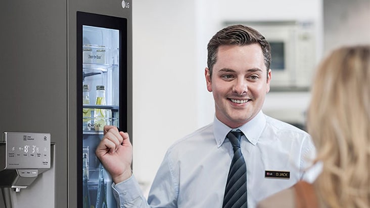 A man in a shirt and tie smiling while indicating a refrigerator behind him as he speaks to a woman with her back to the camera.