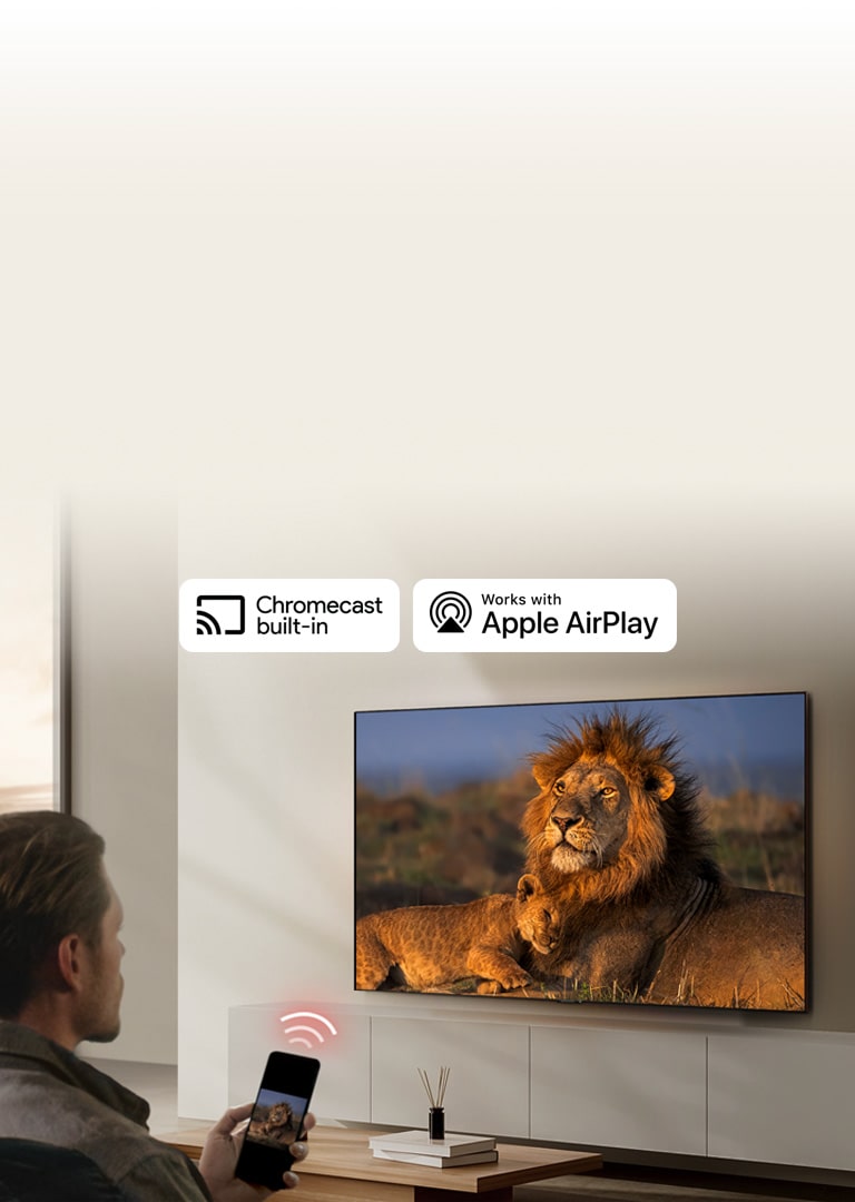  An LG TV mounted on a wall in a living room, displaying a lion and lion cub. A man sits in the foreground with a smartphone in his hand displaying the same image of lions. A graphic of three neon red curved bars is displayed just above the smartphone pointing toward the TV.