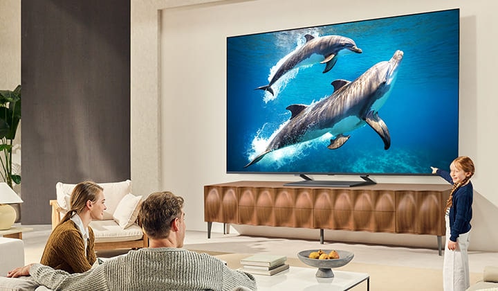 Una niña y un perro están sentados frente a un televisor LG QNED montado en una pared que muestra tres elefantes caminando hacia el exterior sobre una barra de sonido LG. 