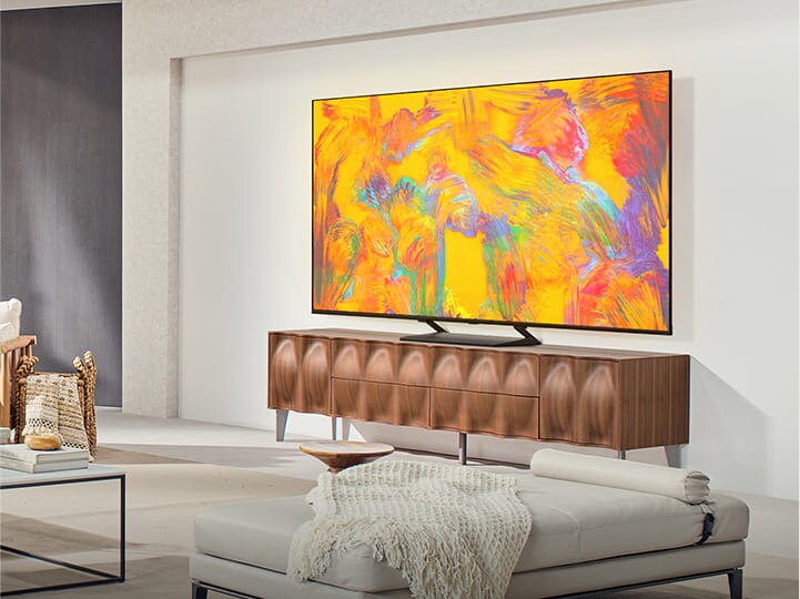 LG QNED TV standing above a TV drawer in a cozy living space. LG QNED TV depicts an abstract background in orange, yellow, purple and pink.