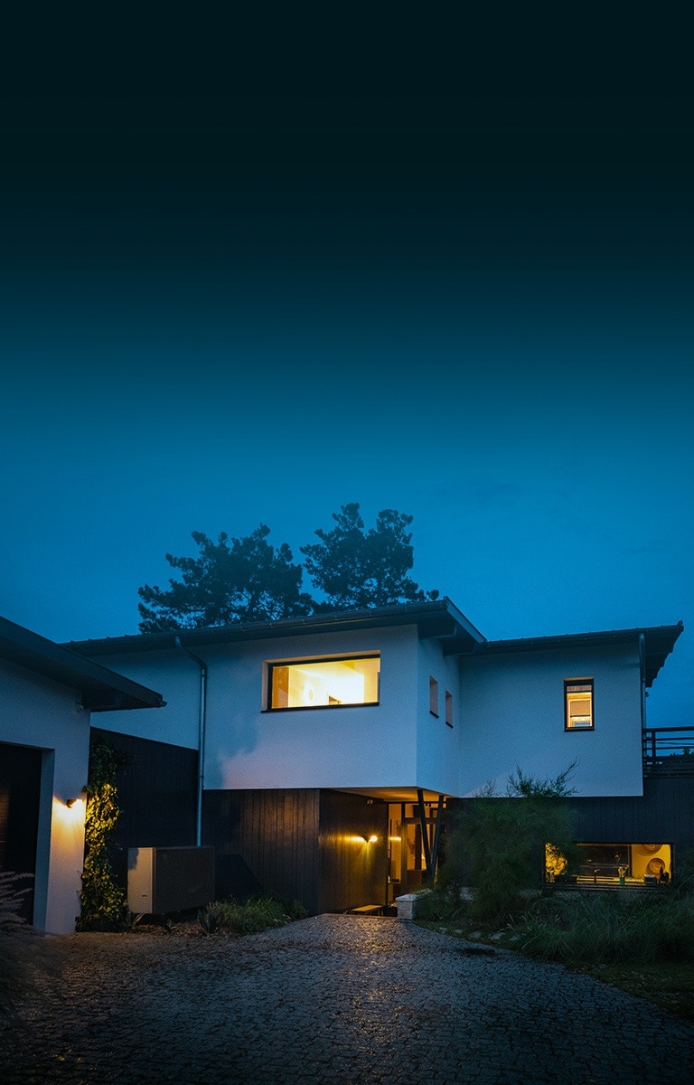 En una noche fría de invierno, plano general del exterior de la casa suavemente iluminada en el interior. Colocación de la nueva bomba de aire-agua negra THERMA V de LG en frente de la casa. 