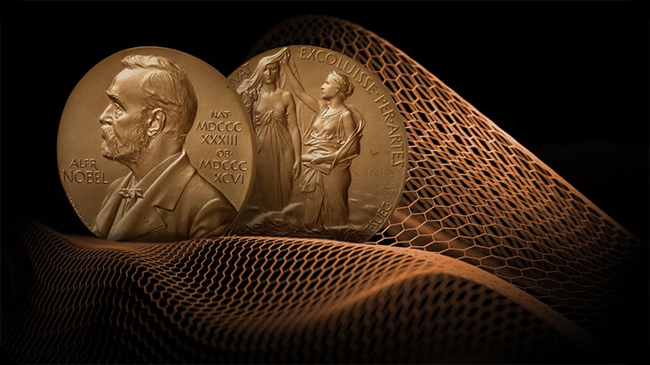 La imagen muestra el anverso y el reverso de la medalla del Premio Nobel. Detrás de la medalla, un dibujo hexagonal en forma de red la rodea para ilustrar que el material grafeno ha ganado el Premio Nobel de Física.