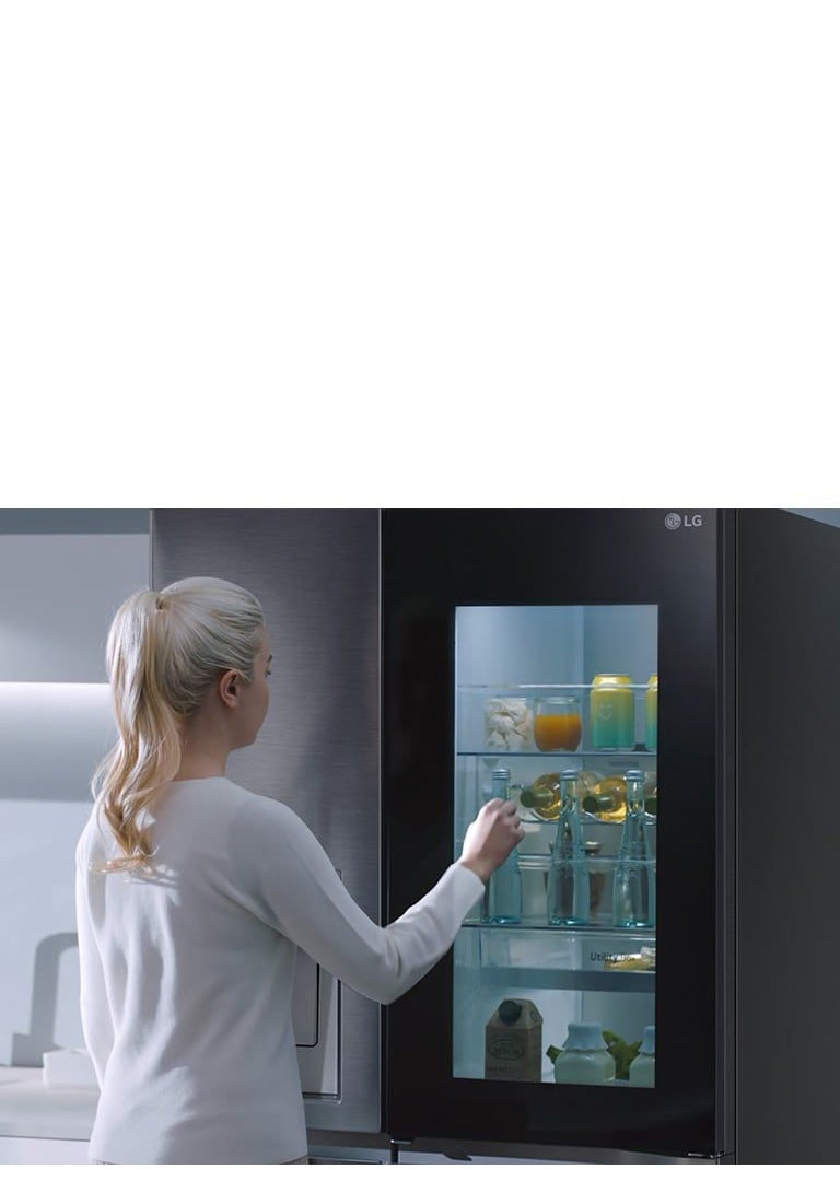 Mujer rubia frente al frigorífico LG, la puerta de cristal revela bebidas y alimentos en el interior.