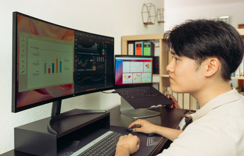 Hombre trabajando en un escritorio moderno con monitores amplios y un portátil en un soporte, enfocado en gráficos y datos en las pantallas.