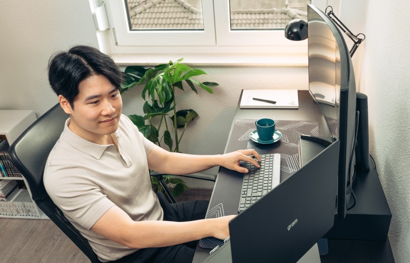 Hombre sentado en un escritorio en una oficina en casa luminosa llena de plantas, trabajando en un portátil LG gram conectado a un monitor externo.