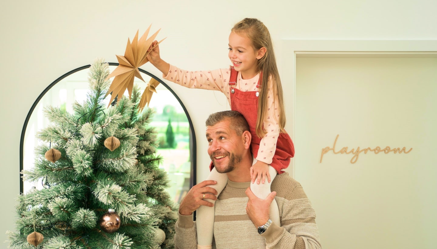 Un padre levanta a su hija sonriente sobre los hombros mientras decora el árbol de Navidad. Cerca se ve una puerta con el letrero 'sala de juegos'.