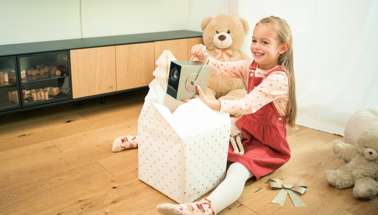 Una niña sonriente abre un regalo de Navidad y descubre un proyector LG CineBeam. Está sentada en un suelo de madera.