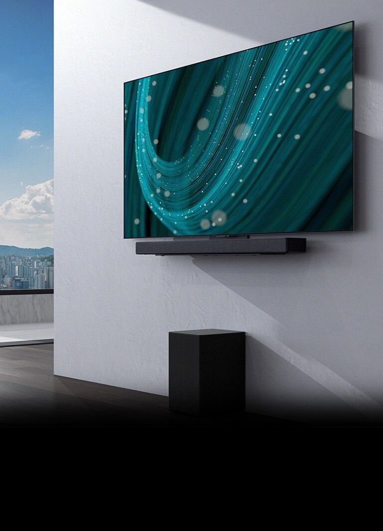 En el centro de un espacio con un gran ventanal, hay una pared con un televisor y una barra de sonido instalados en ella, y un subwoofer debajo. La pantalla muestra una imagen de fondo de color verde azulado.