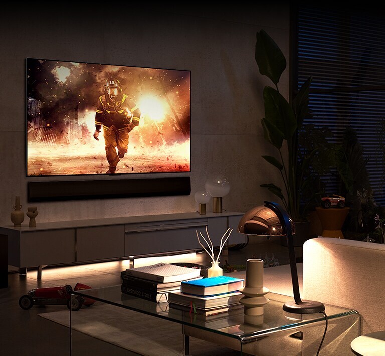 En un salón amplio y cómodo, hay un televisor y una barra de sonido instalados en la pared. En la pantalla del televisor se ve a un bombero saltando de un edificio en llamas.