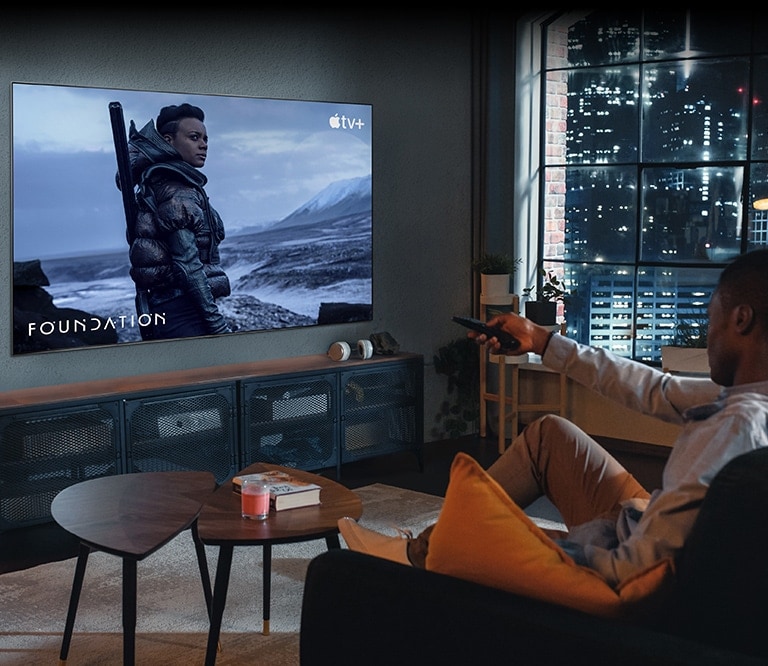 Un hombre está sentado en el sofá viendo la televisión. El hombre tiene un mando a distancia en su mano y, en el TV, se ve la imagen de una escena de Foundation de Apple TV+.