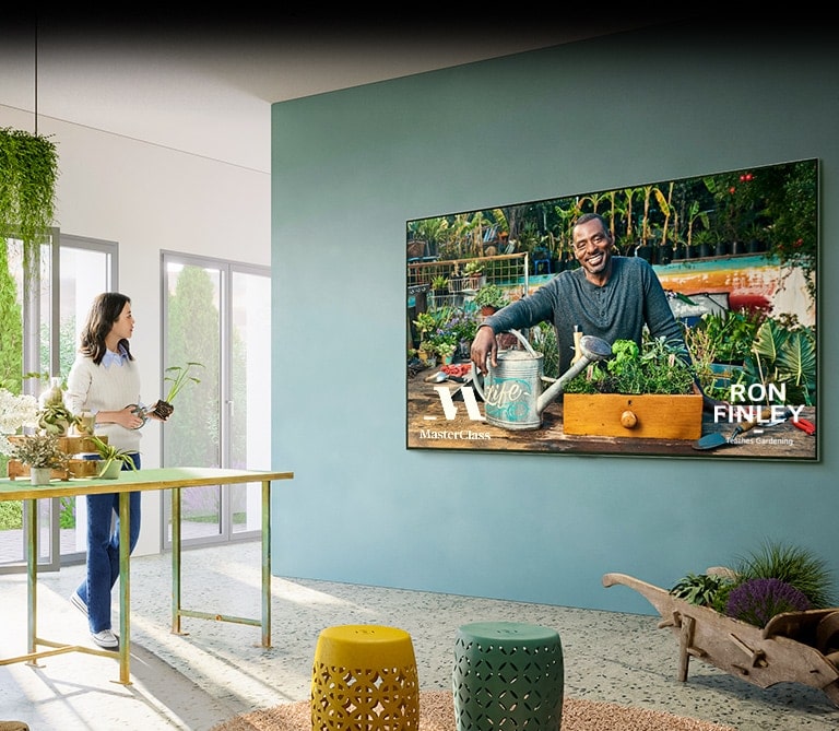 Hay un TV grande en la pared y muestra una clase de jardinería de «Master Class». Una mujer está sentada en una mesa junto a un TV con macetas y tijeras de podar, siguiendo una clase de jardinería.