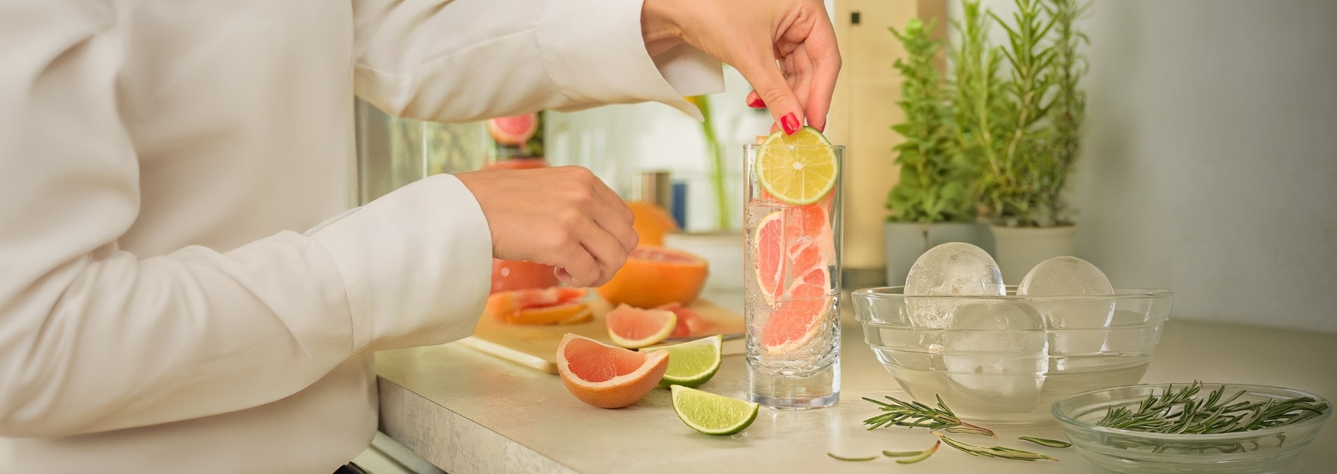 Une femme met des pamplemousses et un citron vert dans un grand verre, des fruits, des herbes et un bol de glaçons sont éparpillés autour du verre.