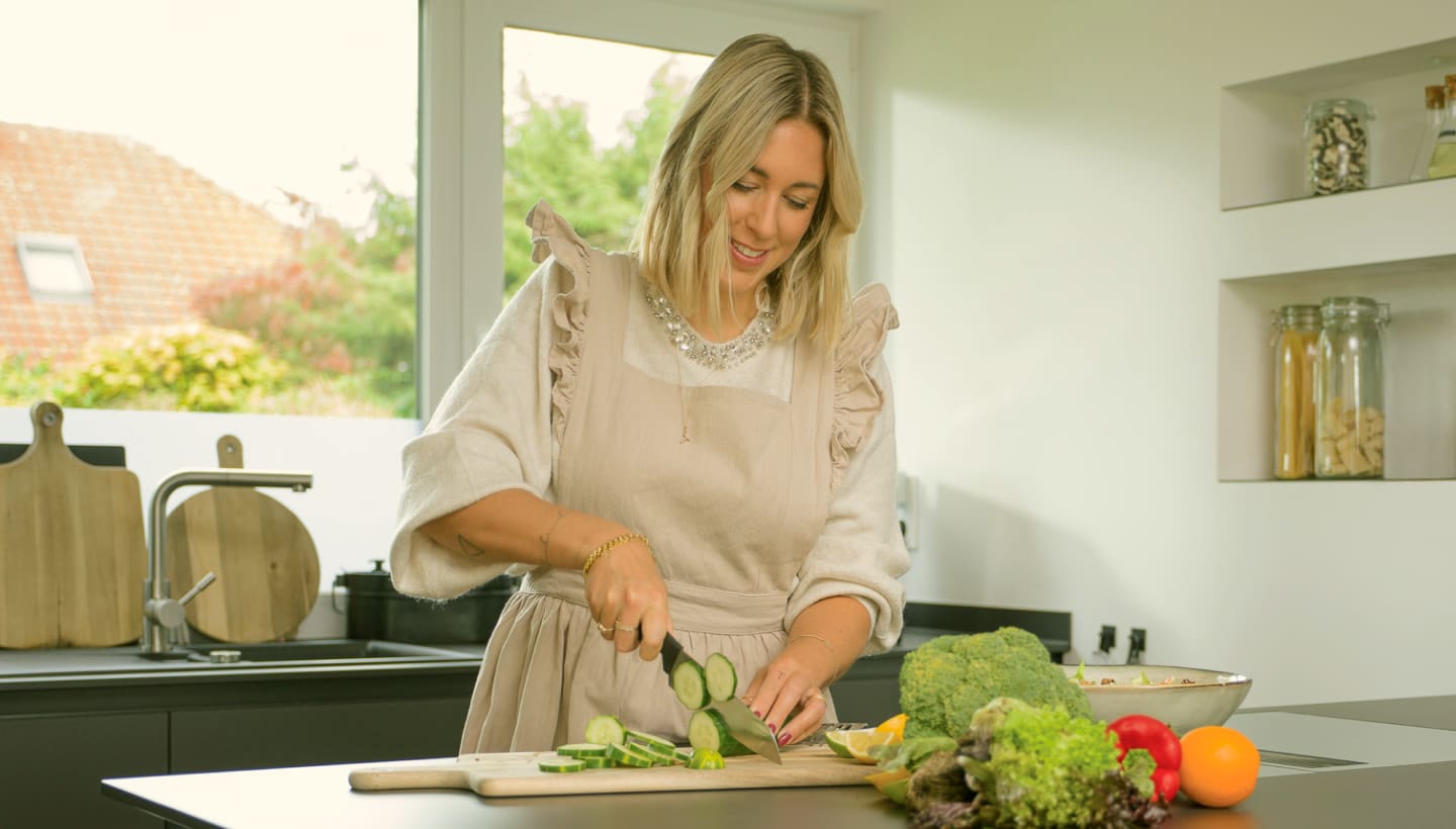 Une femme coupe des concombres dans une cuisine lumineuse avec des légumes colorés sur le plan de travail.