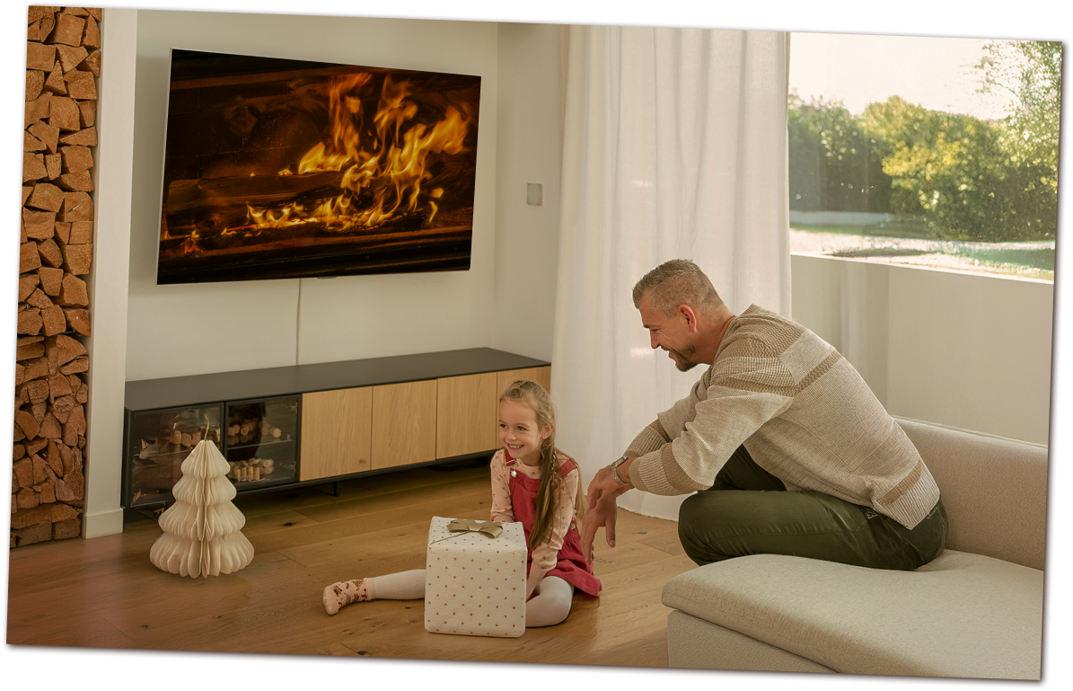 Un père et sa fille célèbrent Noël près d'un LG OLED TV affichant un feu de cheminée. La fille tient un cadeau, entourée d'une décoration conviviale.