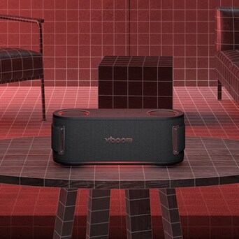 LG XBOOM speaker placed on a table in a red-toned room with grid-patterned walls and modern furniture