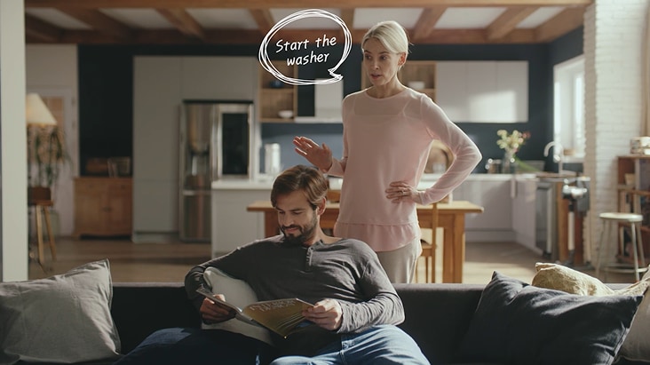In a modern smart kitchen, a woman uses a voice command to start the washer with LG ThinQ AI, while a man reads on the sofa in the background.