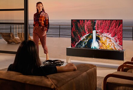 A woman sits on a couch watching a vivid nature scene on a Rollable OLED TV R while another woman stands next to it.