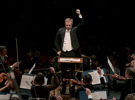 A conductor directs an orchestra. Overlayed text: 'The Musical Offering to the Whole World. #music #partnership'