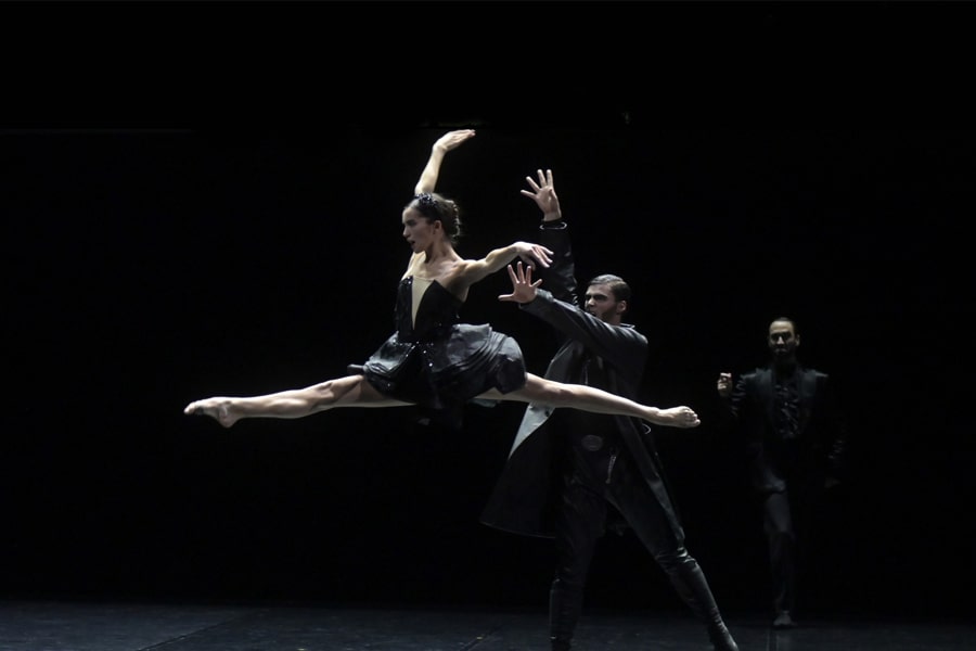 All in black costumes, a female dancer does the splits mid-air, with two other male dancers standing further back.