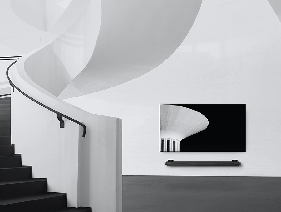 a black and white image of lg signature oled tv which is hung on the wall and to the left there is a spiral staircase