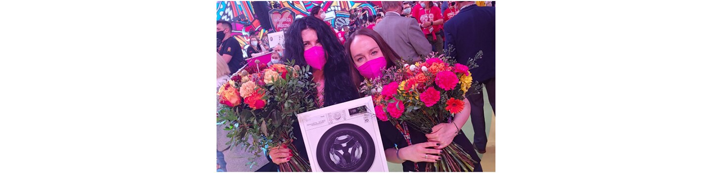 Two female fundraisers holding flowers and a picture of the donated LG Vivace washer dryer in the studio.