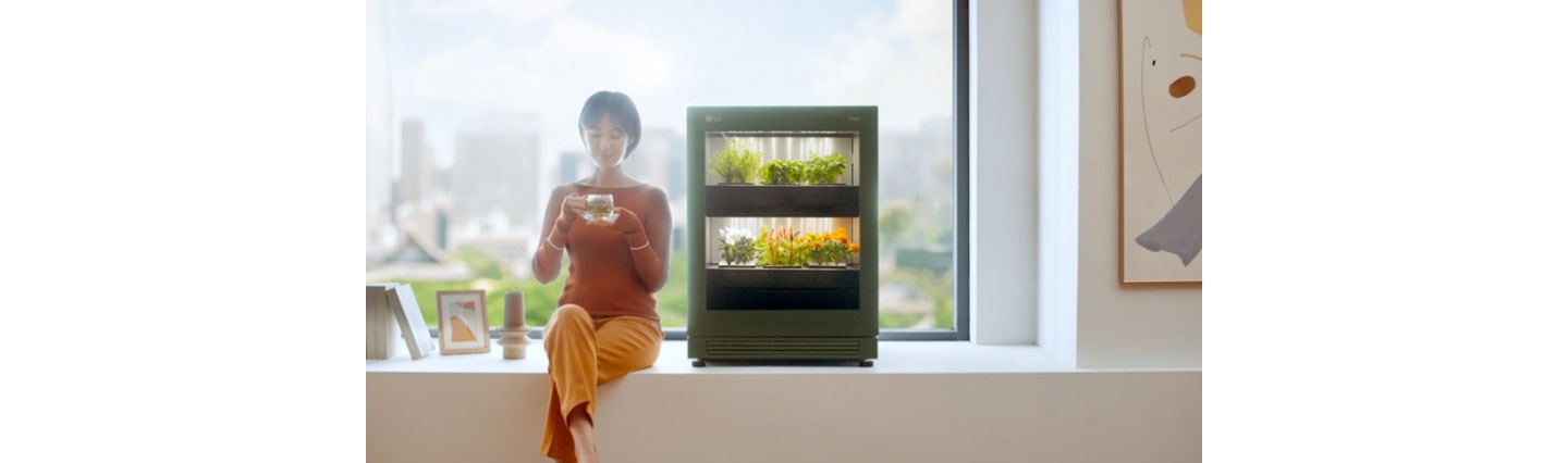 A woman sitting on a large windowsill that looks out on the city as she enjoys a cup of tea next to LG tiiun.
