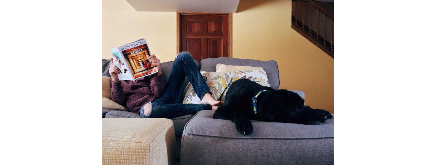 A Man sits on his couch with his dog while reading a magazine.