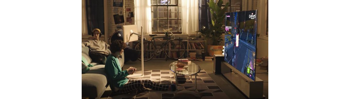 One friend plays a video game on LG’s latest OLED TV while the other lays on the living room sofa next to LG StanbyME.