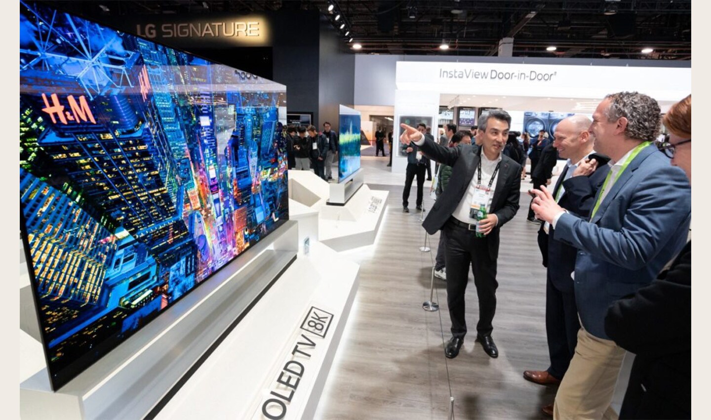 Side view of the display zone of LG's 8K OLED TV, the TV is to the left and a group of attendees on the right admire its excellent features.