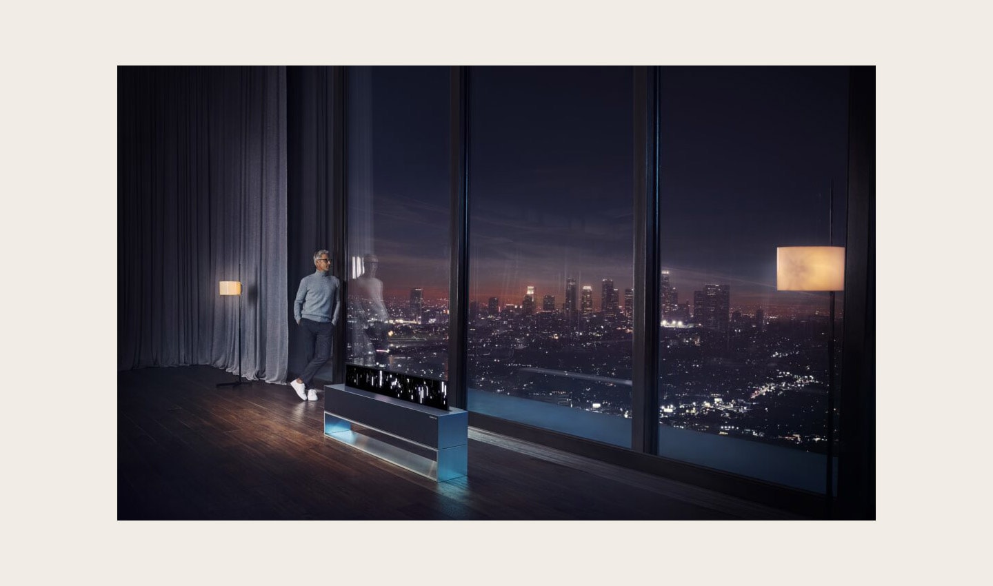 A man looking out the window at the city skyline at night, an LG SIGNATURE OLED TV R is reflecting a picture of the skyline