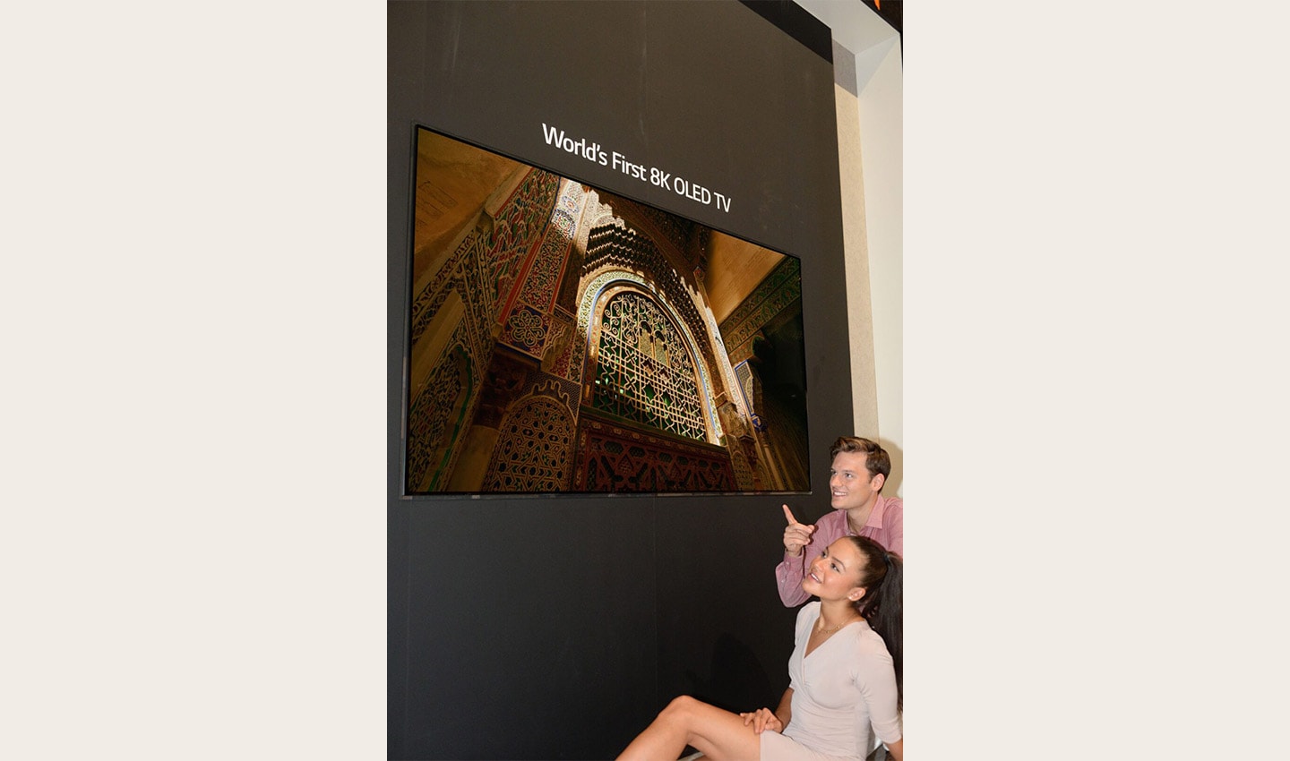 Another view of the World’s First 8K OLED TV display at IFA 2018, with a male and female model sitting below and pointing up to the screen