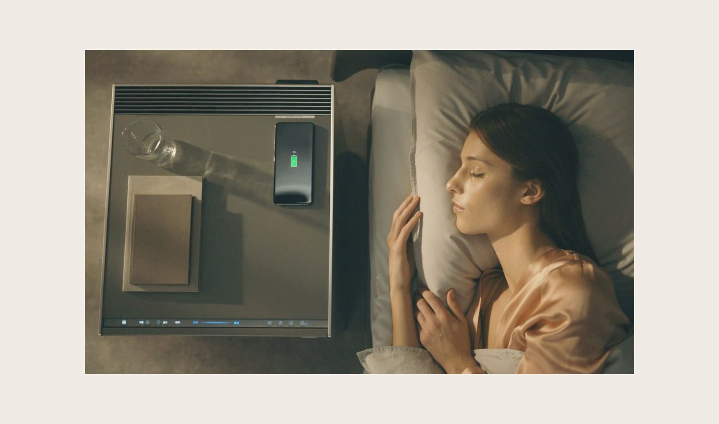 A woman sleeps with an LG OBJET Air Purifier next to her bed; a glass of water, smartphone and books have been placed on top of the purifier