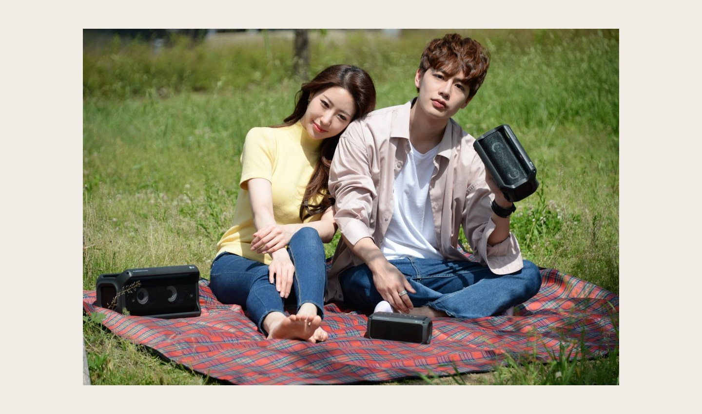 A man and a woman sitting outside on a picnic blanket enjoying music on their LG XBOOMs