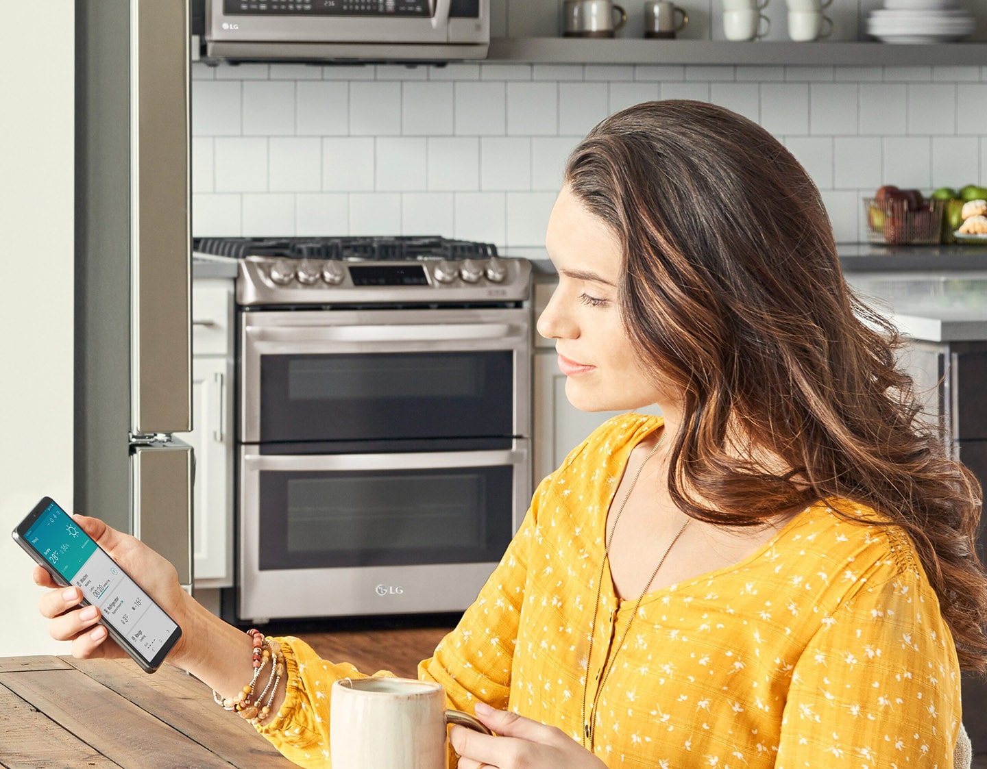 Woman sitting in her kitchen looking at a phone displaying the ThinQ™ app screen showing the device status of the washer, refrigerator and range