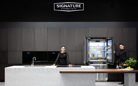 A woman is standing behind the kitchen island and a man is holding the Signature Kitchen Suite built-in 36-inch French Door refrigerator door at LG booth