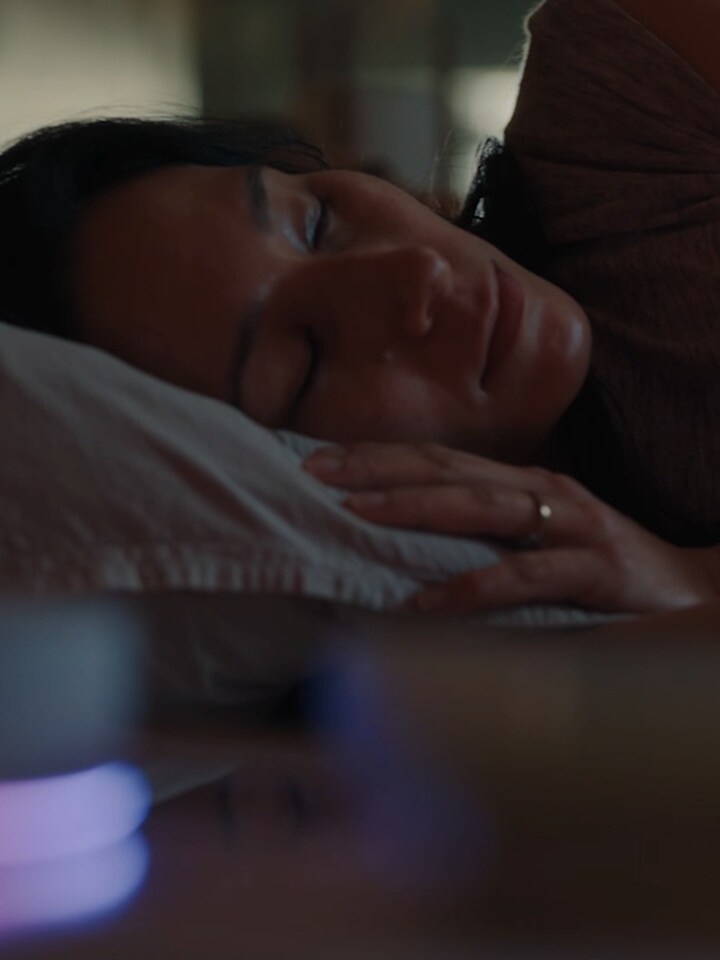 A woman lies on her side, sleeping in bed. In front of her, the LG ThinQ ON device is glowing. 