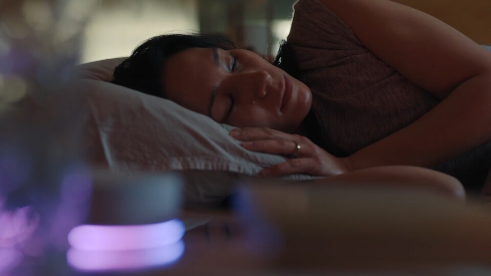 A woman lies on her side, sleeping in bed. In front of her, the LG ThinQ ON device is glowing. The curtain opens, and a man strokes a dog. The woman who was lying down wakes up.
