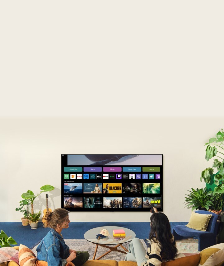 A whale floating above the ocean with a woman in the ocean are shown. A home screen appears from below. As the scene transitions, it shows the image in a large LG TV on the wall. Two women sit in a cozy and neutral living room filled with plants, and a guitar. One woman points the remote at the TV, which shows a range of apps and recommended TV shows on the home screen. 