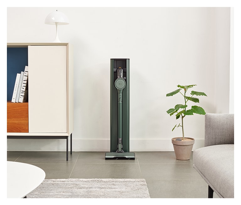 LG vacuum cleaner docked in a station between a white cabinet with books and a potted plant in a modern room.